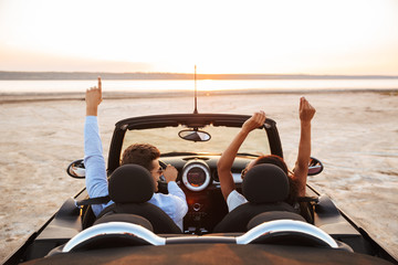 Image from back of young multiethnic couple rejoicing while driving convertible stylish car by...