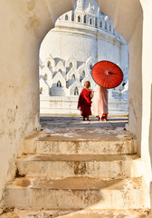 novice buddhist monks with red traditional robes holding red umbrellas walking in a white buddhist temple in myanmar