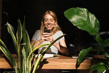 Portrait of blonde young woman smiling and holding smartphone while sitting in cafe