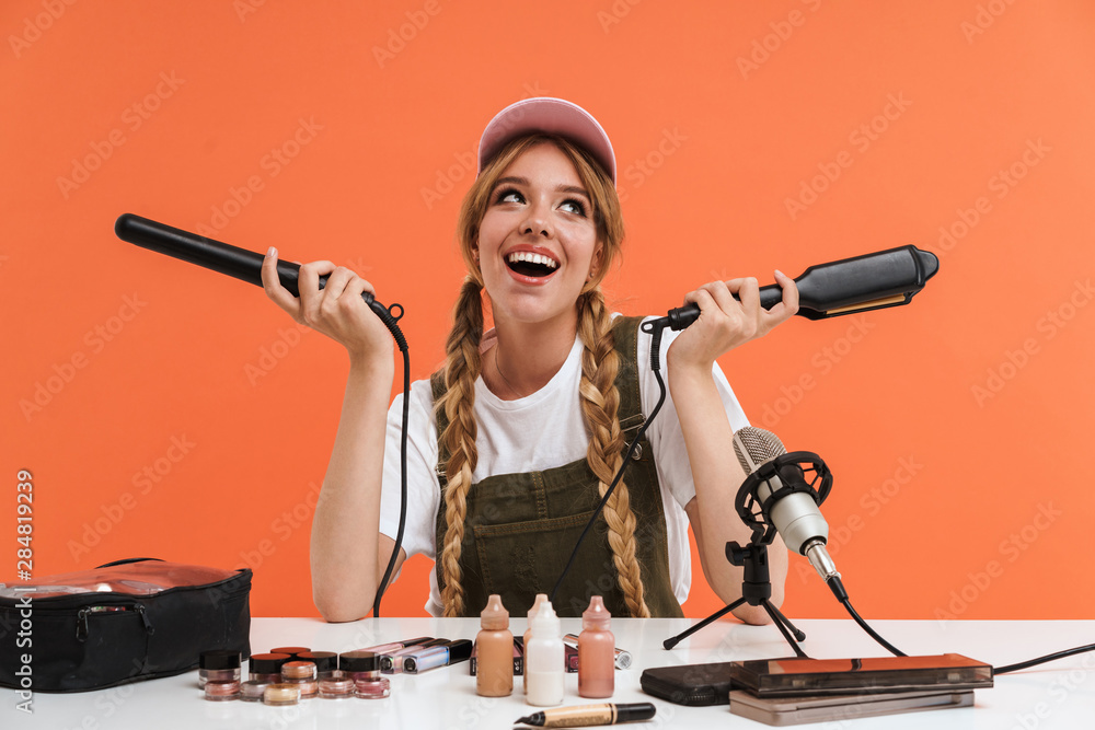 Wall mural Image of young girl holding hair straighteners while recording blog broadcast about new cosmetic products