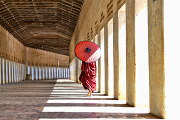novice buddhist monks with red traditional robes holding red umbrellas walking in a white buddhist temple in myanmar