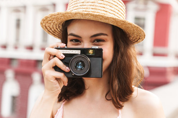Happy young pretty woman in beautiful hat walking by street holding camera.