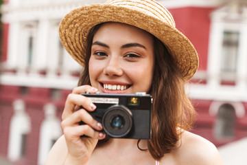 Happy young pretty woman in beautiful hat walking by street holding camera.