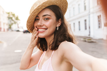 Cheerful young pretty woman in beautiful hat walking by street take selfie by camera.