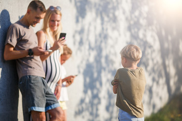 Image of young parents, girl with phones in hands and boy next to gray concrete wall outside on summer day