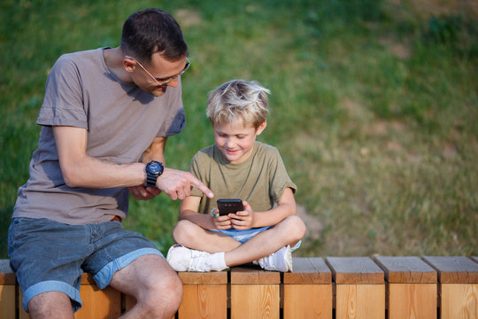 Image of happy man and boy with phones in hands sitting behind wooden fence in park