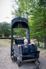 Outdoor portrait of young child sitting in an old black iron mining carriage wagon wearing a white hard hat.