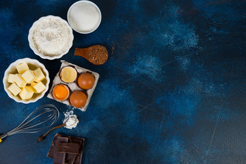A set of basic ingredients for making brownie chocolate cake on a blue concrete background. Chocolate, flour, sugar, eggs, butter and cocoa are on the table. Horizontal orientation with copy space