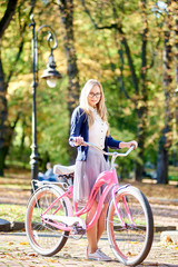 Obraz premium Young happy attractive blond long-haired woman in glasses, skirt and blouse standing with pink lady bicycle on sunny paved park alley on blurred golden yellow green bokeh foliage background.