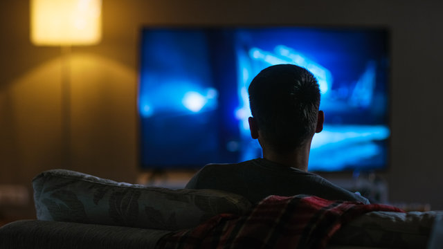 Back View of a Man Sitting on a Couch Watching Movie on His Big Flat Screen TV. 
