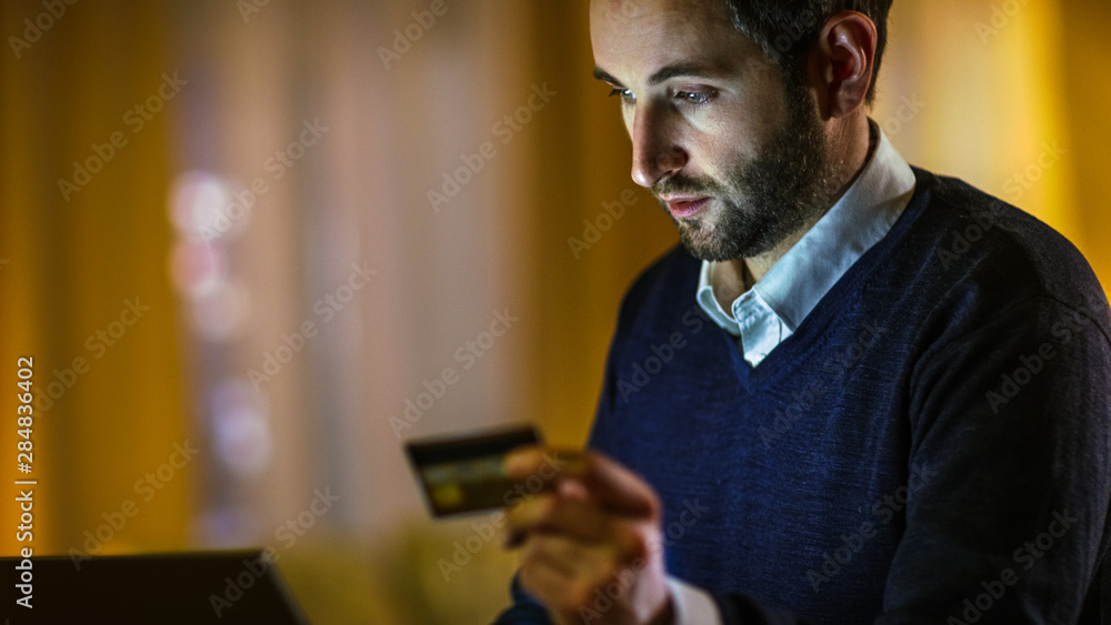 Poster close-up of a man at home he holds credit card and types it's number into his laptop. he's paying fo