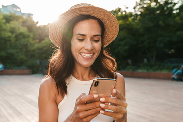 Image of pretty adult woman typing on cellphone and smiling while walking in summer park