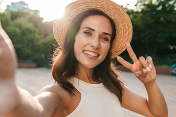 Image of cute middle-aged woman taking selfie photo and gesturing peace sign while walking in...