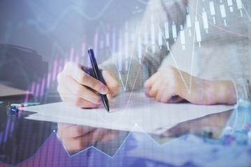 Hands writing information about stock market in notepad. Forex chart holograms in front. Concept of research. Multi exposure