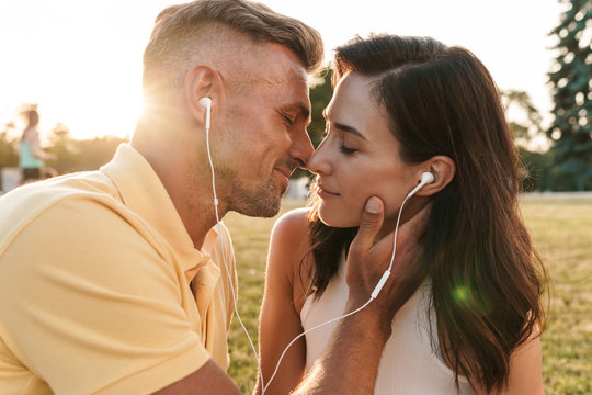Portrait of romantic middle-aged couple man and woman kissing and listening to music together with earphones in summer park