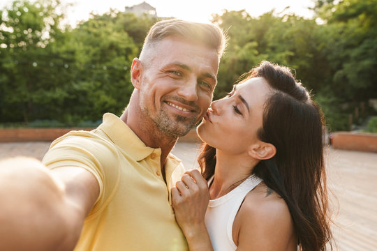 Portrait of pleased middle-aged couple taking selfie photo and kissing while walking in summer park