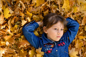 Beautiful little girl in denim shirt is lying on yellow leaves on warm and sunny autumn day. Happy childhood concept