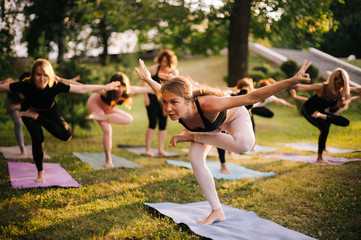 Group of young women are doing difficult yoga exercise standing on one leg. Group of people are straightening her hands in city park on summer sunny morning under guidance of coach.