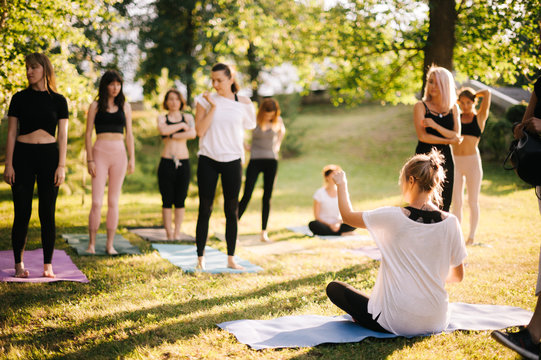 Yoga woman coach welcomes his students and gives instructions in park at dawn. Guru sits on yoga mat among his followers. Beginning of retreat outdoors in sunny morning. Women friendly community