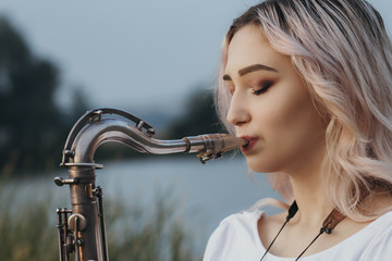 young woman face profile playing saxophone on bank of the river, female blowing into the trumpet, on nature background, concept music and relax - obrazy, fototapety, plakaty