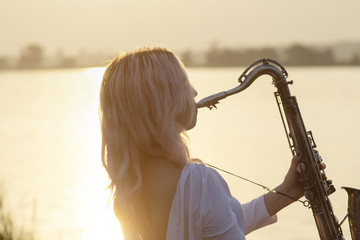 silhouette of a young beautiful girl playing the saxophone at sunrise by the river, woman on the...