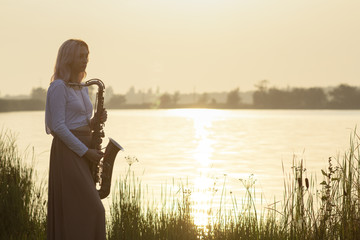 silhouette of a young beautiful girl playing the saxophone at sunrise by the river, a woman in a...