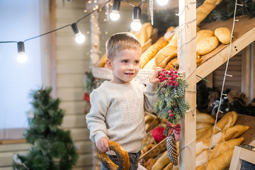 A little boy in a knitted white sweater stands on a stepladder at a bakery, chooses pastries, laughs. The showcase is decorated with Christmas trees, new year garlands, snow and icicles. Fresh bread 