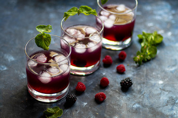 Blackberry tincture in a transparent glass. Ice cubes and pieces of fruit are also in the middle of the dishes. All on a dark background. Slices of lemon and mint leaves serve. 