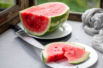 A slice of watermelon in the foreground. In the background is half a berry on a white plate. Near a big knife. 