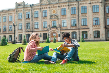 Smiling African American student with glasses and with books and girl have fun chatting near college. Happy friends are talking on the background of the university. 