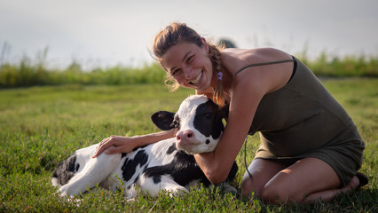 Authentic shot of young woman farmer is caressing  an ecologically grown newborn calf used for biological milk products industry on a green lawn of a countryside farm with a sun shining. © Kitreel