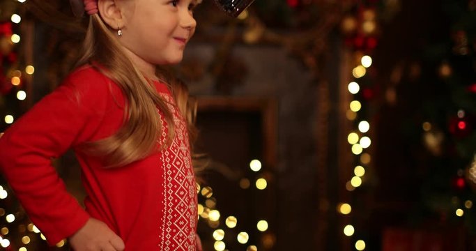 A Pretty Little Girl Takes Off The Christmas Cap In Apartment With Christmas Lights And Decorations. Merry Christmas, Happy New Year. Miracle Time.