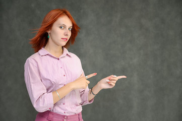 Photo Portrait of a cute female girl manager with bright red hair manager in a pink shirt on a gray background in the studio. He talks, shows his hands in front of the camera with emotions.