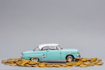 Light blue car figurine placed on top of big pile of coins facing towards the right, on gray background.