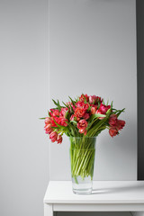 Beautiful bouquet of red tulips in a glass vase on a white table in a bright interior . Against a light gray wall.