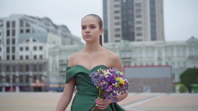 Beautiful girl in chic dress standing with fragrant bouquet of wildflowers on the background of the morning city. Real people series.