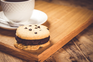 Cookie butter and chocolate Cookies stacked 3 pieces, black coffee in the cup white on white coasters,and stainless steel spoon are on a wooden tray ,  all placed on a wooden table.