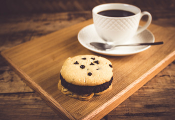 Cookie butter and chocolate Cookies stacked 3 pieces, black coffee in the cup white on white coasters,and stainless steel spoon are on a wooden tray ,  all placed on a wooden table.
