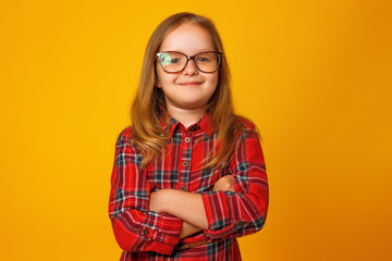 Portrait of a little blonde girl in a red dress on a yellow background. The child is standing in glasses with arms crossed.