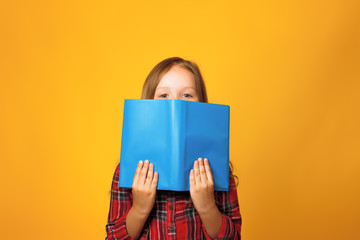 Cheerful little girl student on a yellow background. The child is hiding behind an open book and...