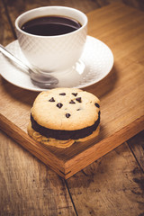 Cookie butter and chocolate Cookies stacked 3 pieces, black coffee in the cup white on white coasters,and stainless steel spoon are on a wooden tray ,  all placed on a wooden table.