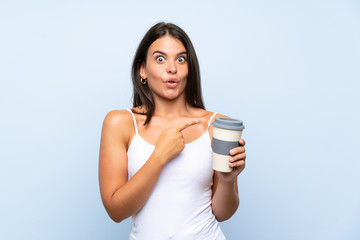 Young woman holding a take away coffee over isolated blue background surprised and pointing side