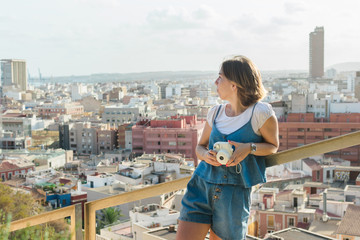 Young woman is shooting with a instant camera with the city of Alicante on background