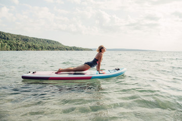 Middle age Caucasian woman practising yoga on paddle sup surfboard at sunset. Female stretching...