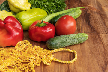 Green, yellow and red vegetables lie next to a yellow string bag on a brown wooden surface, side view from above