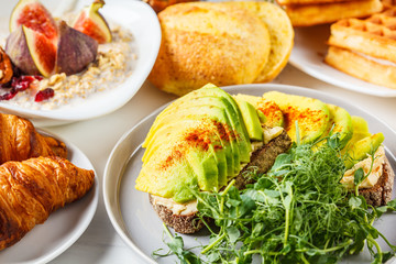 Breakfast table with avocado toast, oatmeal, waffles, croissants on white background.