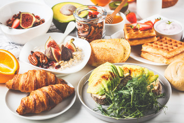 Breakfast table with avocado toast, oatmeal, waffles, croissants on white background.