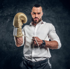Cheeky handsome man in white shirt and with golden boxing glove is standing at dark studio.