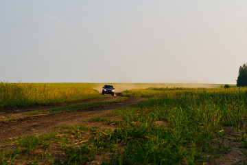 Red crossover raises a cloud of dust on a country dirt road. The road along the green meadow.