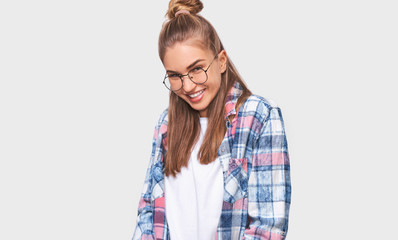 Satisfied young woman dressed in casual plaid shirt, wearing round transparent eyewear with pleasant broadly smile, looking to the camera and posing over white studio wall. People emotions